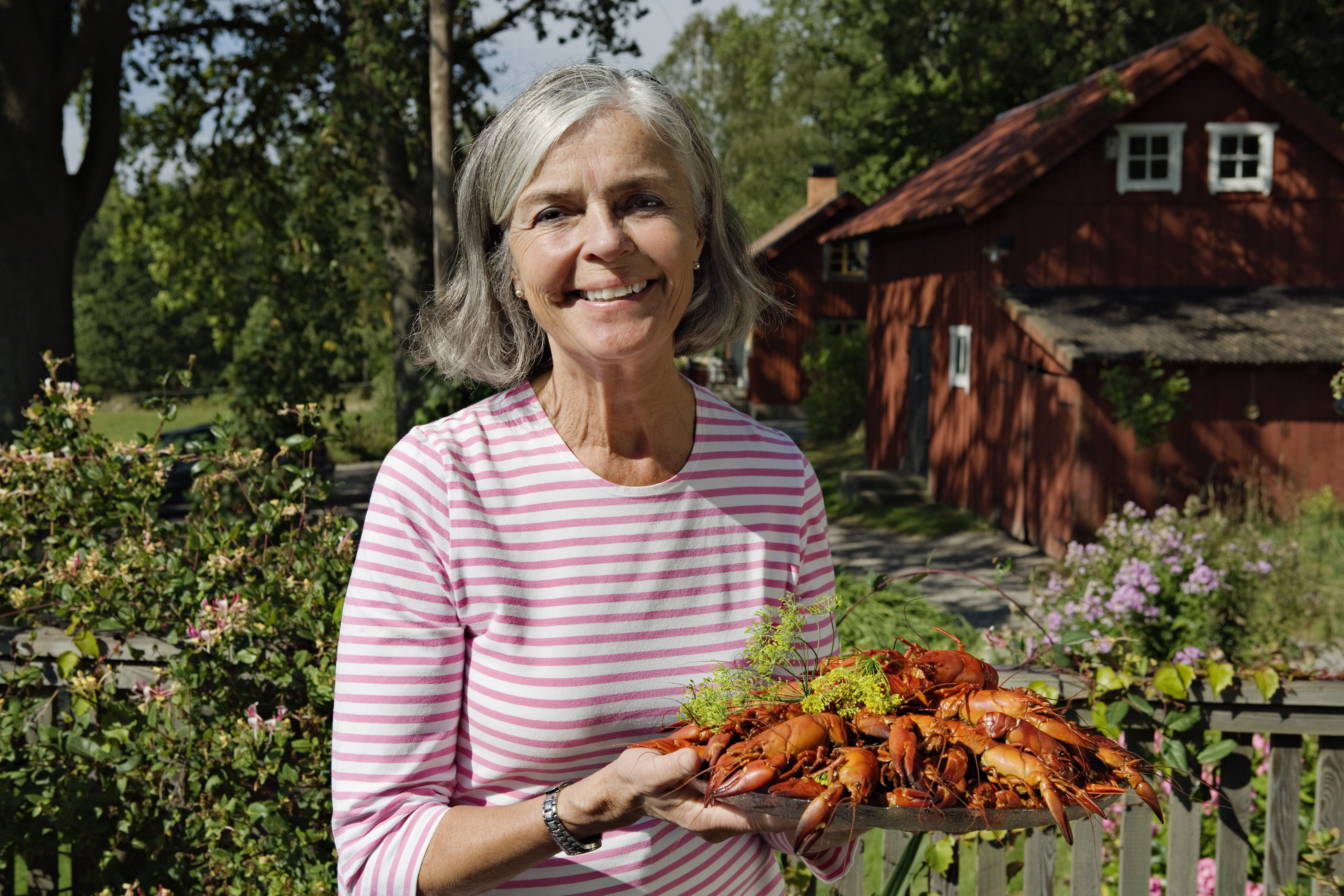 En äldre kvinna står i en trädgård och håller ett fat med kräftor